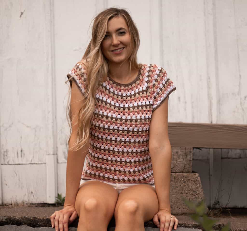 maria top crochet pattern main image, model sitting on sidewalk in front of white building wearing a multicolored crocheted top worked in granny stitch