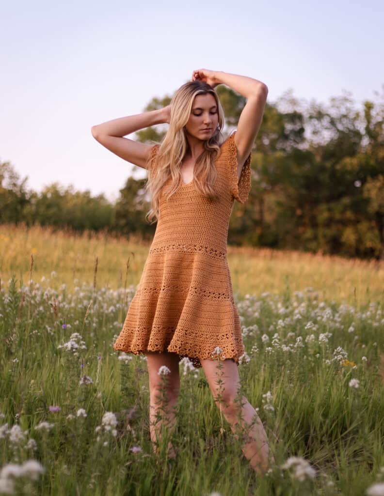 sun dress being worn on model standing in field dancing with her arms up. The dress is a butterscotch color and is flowy at the waist with lace flutter sleeves