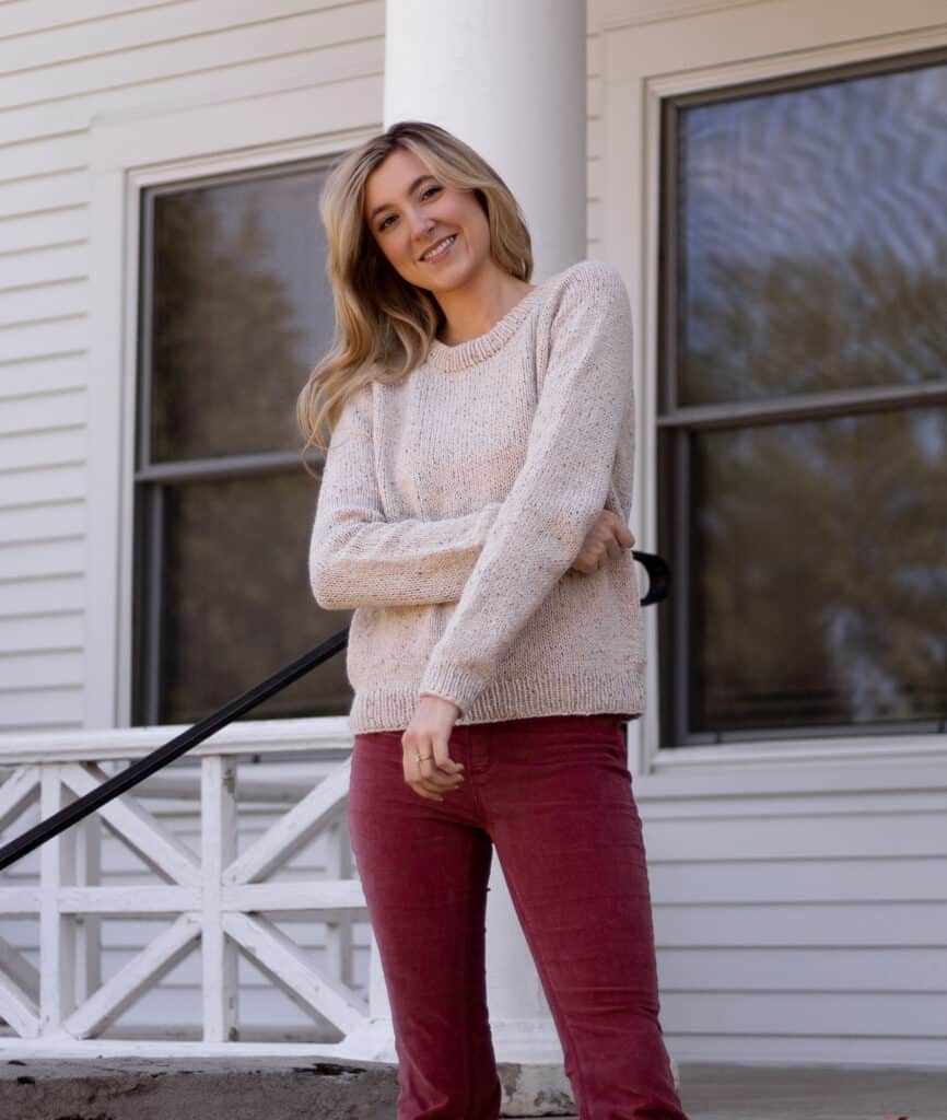 Girl wearing a hand knit top down raglan style sweater with her arms crossed in front of her smiling at the camera. The sweater is tweed and looks textured from the yarn