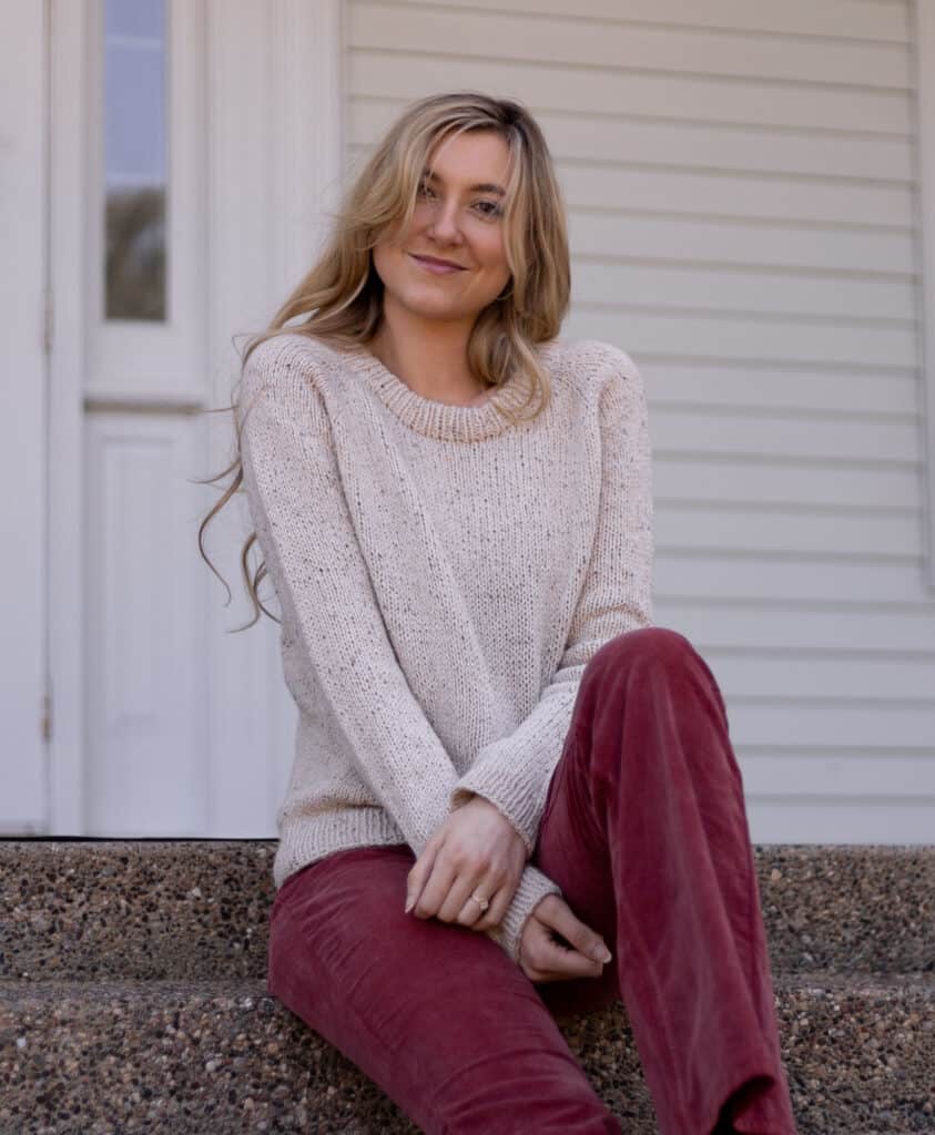 Model sitting on steps smiling swearing a tan, tweed hand knit sweater.  The sweater is classic and looks comfortable