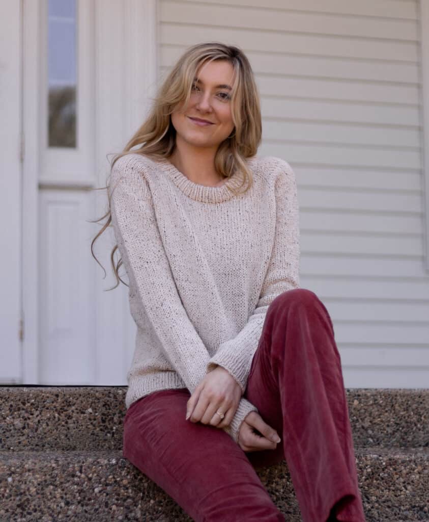 Model sitting on steps smiling swearing a tan, tweed hand knit sweater. The sweater is classic and looks comfortable