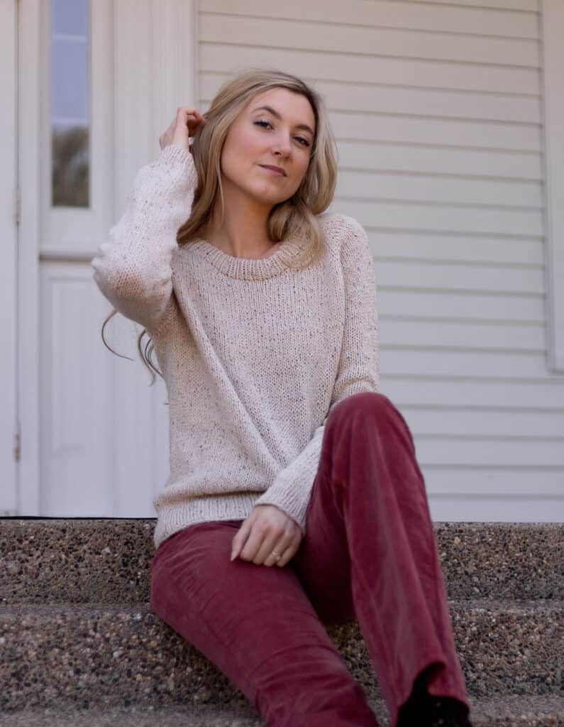 Woman wearing a tan sweater while sitting down. She is smiling slightly at the camera with one hand up. Hand Knit raglan style sweater looks comfortable and cozy.