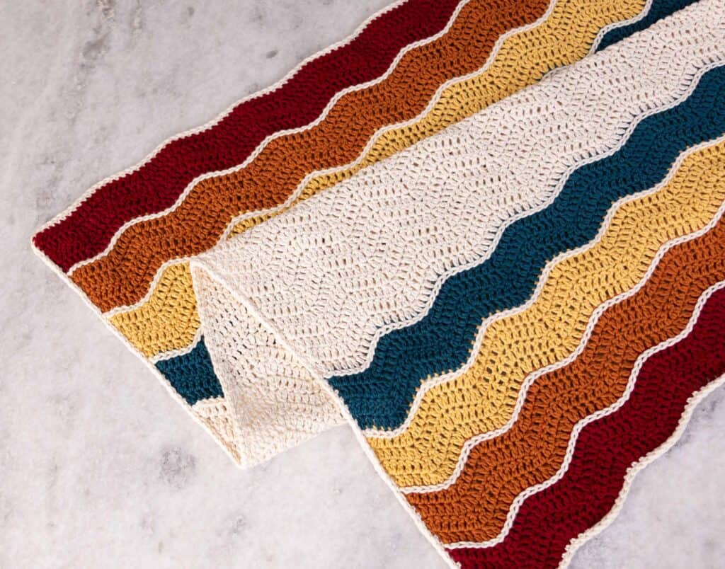 Wavy Baby Blanket draped on top of itself laying on a table. Color detailing is on each edge in the following order: red (Jam), orange (Penny), yellow (banana), and blue (Ocean), with a white main color section between 