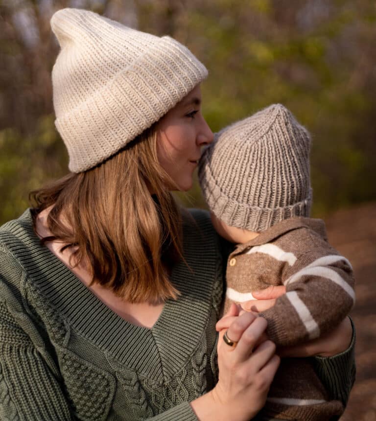 Classic ribbed hats being worn by a mother and her baby. Baby is looking away from the camera and mother kisses the side of the baby's head. Hats are cream and grey with a rib knit stitch and decorative decreases at the crown