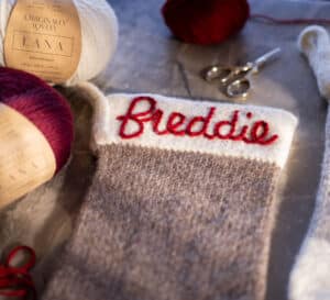 Christmas stocking laying on top of a marble table. Stocking is white and grey with the name 'freddie' embroidered along the top in red. Behind the stocking sits balls of yarn and a pair of scissors.