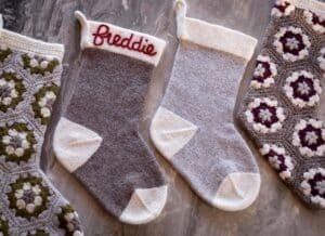 A set of four christmas stockings laying flat upon a grey table. Stockings are knit using a wool yarn and one of them says Freddie along the top in cursive, one is plain, and two are crocheted and multicolored