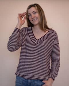 Woman smiling at the camera with one arm lifted wearing the Seasons sweater, a size inclusive knitting pattern made using Originally Lovely Cria yarn. This stripe sweater has drop shoulders and a v neckline