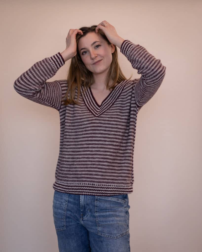 Woman is wearing a striped sweater and putting her hands up in the air on either side of her head. This showcases the positive ease of the sweater and the beautiful drape of the Cria baby alpaca yarn.